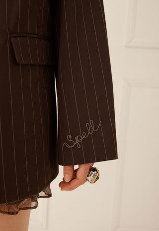 close-up-of-a-dark-brown-pinstripe-blazer-with-thin-white-vertical-stripes-featuring-a-cursive-embroidered-spell-logo-on-the-cuff-and-a-hand-holding-a-ring-near-the-hem