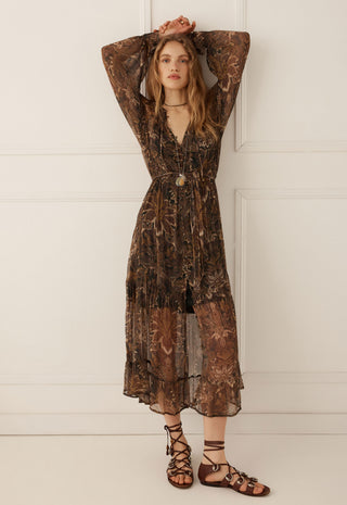 a-woman-modeling-a-long-sleeved-brown-and-black-floral-print-midi-dress-with-a-tie-front-waist-and-a-v-neckline-paired-with-brown-strappy-flat-sandals-standing-against-a-white-paneled-wall