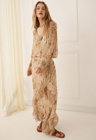 a-woman-modeling-a-long-sleeved-maxi-dress-with-a-beige-and-brown-paisley-pattern-standing-in-a-bright-minimalist-room-with-white-walls-and-a-cream-curtain