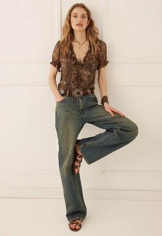 a-woman-posing-in-a-brown-patterned-short-sleeve-blouse-blue-wide-leg-jeans-and-brown-strappy-sandals-against-a-white-paneled-wall