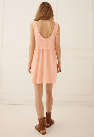 a-woman-viewed-from-behind-wearing-a-peach-colored-sleeveless-dress-with-a-ruffled-back-and-brown-leather-slide-sandals-standing-against-a-white-paneled-wall