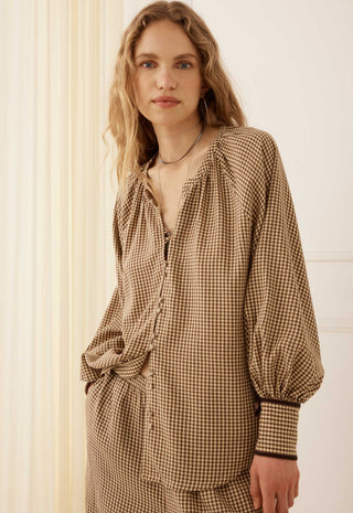 a-woman-wearing-a-brown-and-beige-gingham-long-sleeve-blouse-with-a-gathered-neckline-and-balloon-sleeves-standing-against-a-cream-colored-paneled-wall