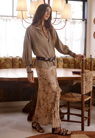 a-woman-stands-in-a-rustic-dining-room-wearing-a-long-sleeve-beige-patterned-blouse-with-a-black-belt-and-a-matching-long-skirt-with-a-paisley-print-she-is-wearing-brown-strappy-sandals-and-leaning-on-a-wooden-chair-next-to-a-table