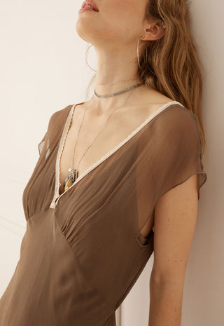 a-close-up-lifestyle-shot-of-a-model-wearing-a-brown-semi-sheer-draped-dress-with-a-deep-v-neckline-and-layered-silver-necklaces