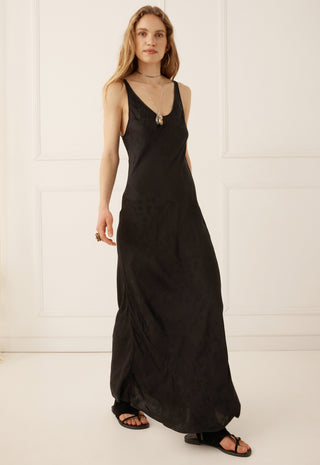 a-woman-modeling-a-black-sleeveless-maxi-dress-with-a-deep-v-neckline-and-a-small-silver-pendant-at-the-neckline-standing-against-a-white-paneled-wall