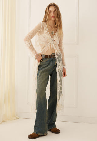 a-woman-stands-indoors-wearing-a-sheer-white-lace-long-sleeve-cardigan-over-a-cropped-top-blue-flared-jeans-and-brown-suede-boots-against-a-white-paneled-wall