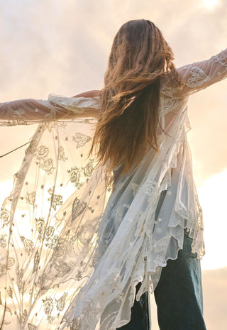 a-woman-seen-from-behind-wearing-a-sheer-white-lace-long-sleeve-duster-or-wrap-with-butterfly-and-floral-embroidery-paired-with-dark-jeans-set-against-a-bright-sunlit-sky