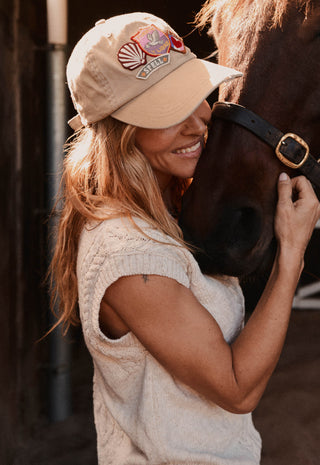 A woman in a cream knit top and beige baseball cap with a horse, smiling in a barn setting. The cap features a colorful embroidered logo on the front.