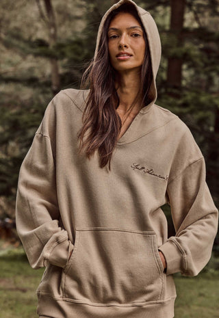 a-woman-wearing-a-beige-oversized-pullover-hoodie-with-a-small-script-logo-on-the-left-chest-photographed-outdoors-against-a-blurred-forest-background