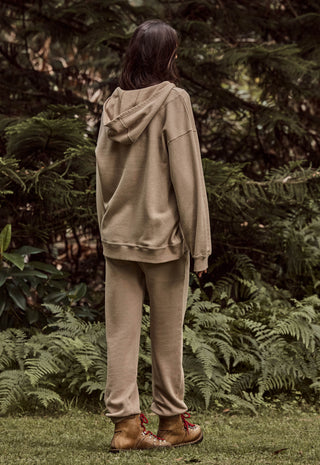 a-person-standing-outdoors-in-a-wooded-area-viewed-from-behind-wearing-a-matching-beige-hooded-sweatshirt-and-jogger-pants-with-brown-lace-up-boots