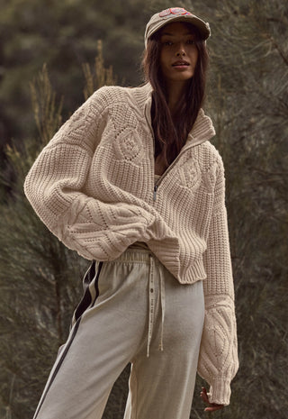 a-woman-wearing-a-chunky-cable-knit-zip-front-sweater-in-a-warm-cream-or-oatmeal-tone-paired-with-light-beige-sweatpants-and-a-beige-cap-standing-outdoors-against-a-blurred-natural-background