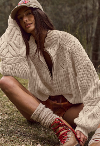 a-woman-crouching-outdoors-in-a-wooded-setting-wearing-a-chunky-cream-cable-knit-half-zip-sweater-a-matching-beige-baseball-cap-brown-plaid-shorts-and-tan-lace-up-boots-with-red-laces