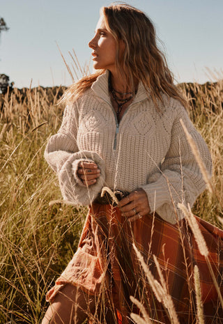 a-woman-standing-in-a-field-of-dry-golden-grass-wearing-a-cream-cable-knit-half-zip-sweater-and-a-rust-orange-plaid-skirt