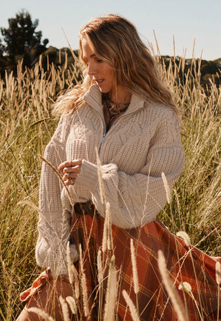 a-woman-standing-in-a-field-of-dry-golden-grass-wearing-a-chunky-cream-cable-knit-quarter-zip-sweater-and-a-rust-orange-skirt