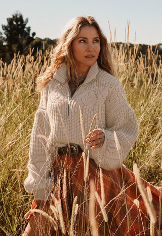 a-woman-standing-in-a-field-of-dry-grass-wearing-a-cream-cable-knit-mock-neck-sweater-and-a-rust-orange-skirt