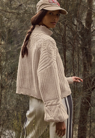 a-woman-wearing-a-chunky-cream-cable-knit-mock-neck-sweater-a-beige-baseball-cap-and-cream-pants-with-black-stripes-standing-in-a-wooded-outdoor-setting