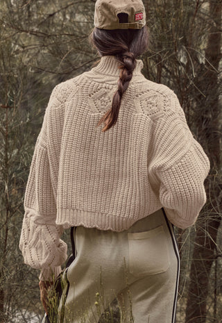 a-person-shown-from-behind-wearing-a-cream-colored-chunky-knit-cropped-sweater-beige-pants-and-a-beige-cap-standing-outdoors-in-a-wooded-setting