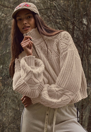 a-woman-wearing-a-chunky-cable-knit-turtleneck-sweater-in-a-warm-cream-beige-color-styled-with-a-matching-beige-baseball-cap-and-beige-sweatpants-set-against-a-blurred-forest-background