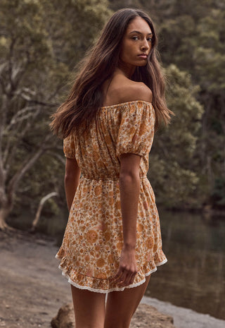 a-woman-wearing-an-off-the-shoulder-mini-dress-with-a-rust-orange-and-beige-floral-pattern-standing-outdoors-near-a-body-of-water-with-trees-in-the-background