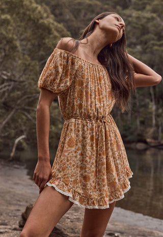 a-woman-wearing-a-short-off-the-shoulder-dress-with-a-rust-orange-and-beige-floral-pattern-standing-outdoors-near-water-and-trees