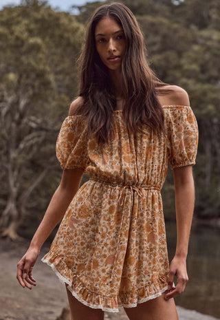 a-woman-wearing-a-short-off-the-shoulder-dress-with-puff-sleeves-and-a-gathered-waist-featuring-an-orange-and-beige-floral-pattern-she-is-standing-outdoors-in-front-of-a-blurred-natural-background-of-trees