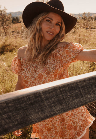 a-woman-outdoors-wearing-a-black-wide-brim-hat-and-a-short-off-the-shoulder-floral-dress-in-orange-and-cream-tones-standing-behind-a-wooden-fence-in-a-sunlit-grassy-field