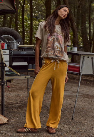 A woman stands outdoors in a campsite setting wearing a beige graphic t-shirt and mustard yellow wide-leg sweatpants. She is wearing brown leather slide sandals.
