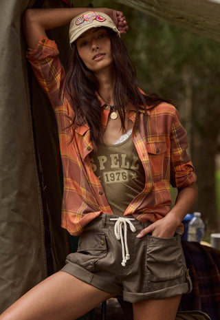 a-woman-posing-in-a-rustic-outdoor-setting-wearing-a-rust-orange-and-yellow-plaid-flannel-shirt-over-a-graphic-tank-top-olive-green-cargo-shorts-and-a-beige-baseball-cap